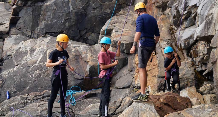 Four people wearing safety gear hold ropes at the base of a rock face